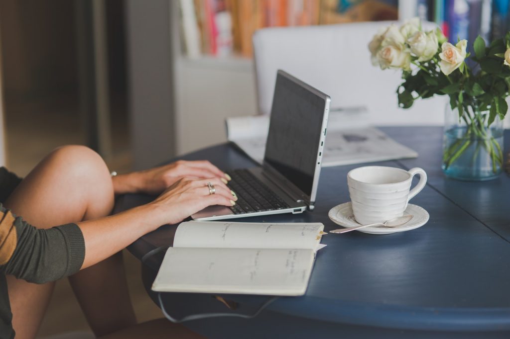 person working at computer on kitchen table with notebook, coffee cup, and vase of flowers nearby 